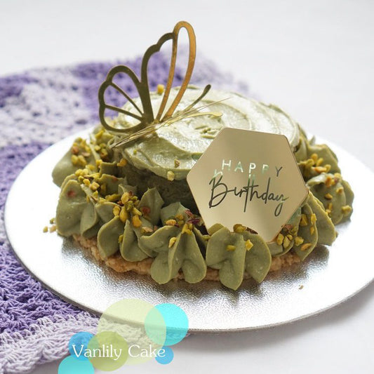 精緻的綠色開心果流心蛋糕，裝飾有金色蝴蝶插件與「Happy Birthday」標牌。蛋糕外層擠滿開心果奶油花並撒上開心果碎，置於紫色編織墊上的銀色底盤，展現低調奢華的健康甜點風格。